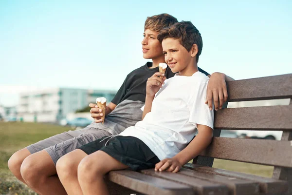 アイスクリームを食べると悲しいことではありません。ビーチのベンチに座っている間にアイスクリームコーンを食べている2人の幸せな兄弟のショット. — ストック写真