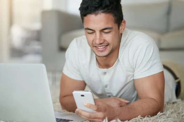 Altijd op de hoogte dankzij moderne technologie. Opname van een ontspannen jongeman met een telefoon en laptop op de vloer thuis. — Stockfoto