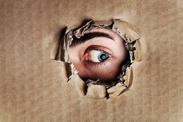 Wat er aan de andere kant is. Close-up portret van een oog kijkend door een gescheurd gat in een stuk karton. — Stockfoto