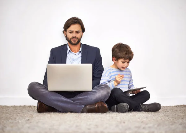 Boys love their digital toys. A father and son sitting on the floor against a wall with a laptop and digital tablet.
