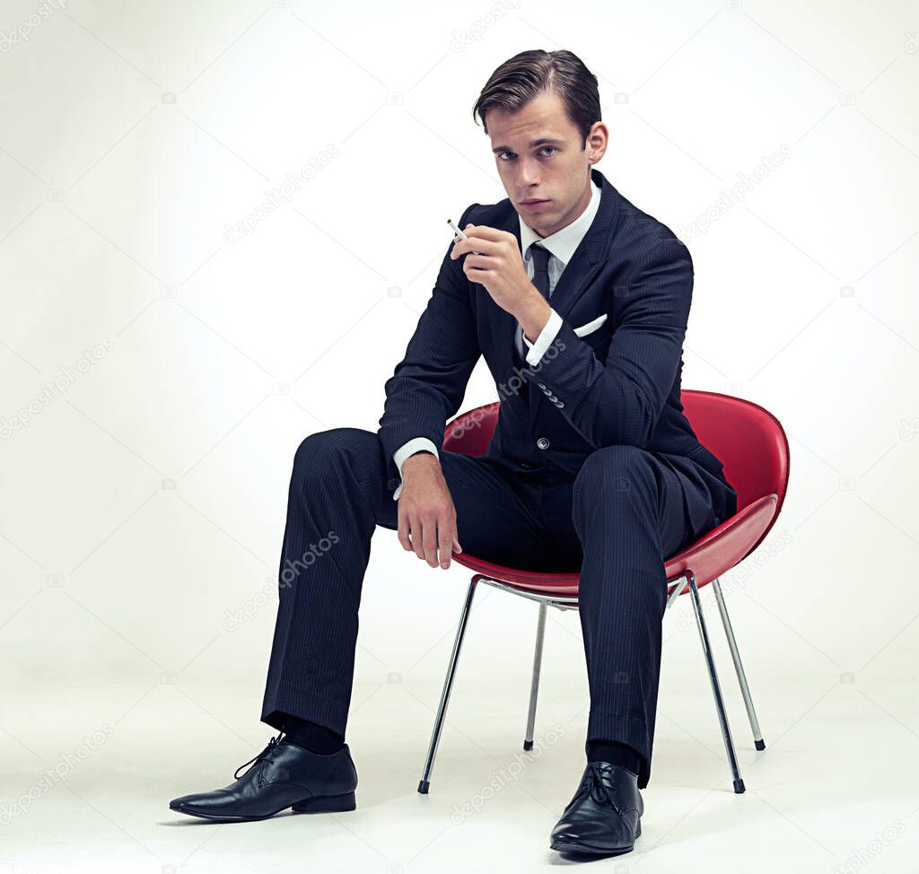 Suave smoker. A studio portrait of a handsome young gentleman in a pinstripe suit sitting on a chair.