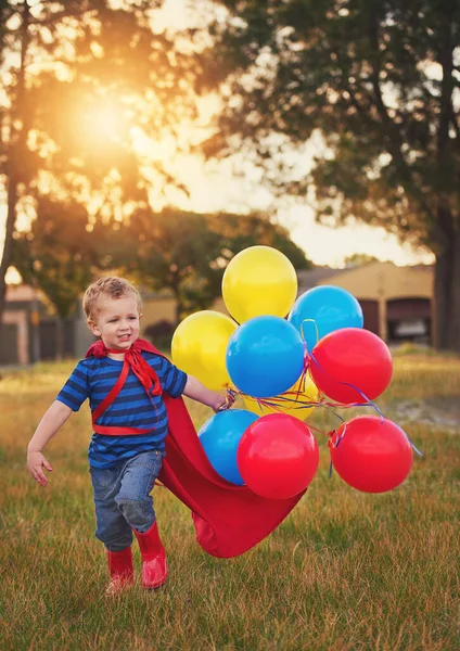 Nächster Halt, der Mond. Aufnahme eines süßen kleinen Jungen im Superheldenkostüm, der einen Haufen Luftballons nach draußen trägt. — Stockfoto