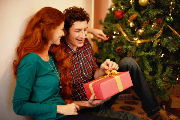 Compartir Navidad con alguien especial. Shot de un joven abriendo su regalo de Navidad mientras se sienta con su novia. — Foto de Stock