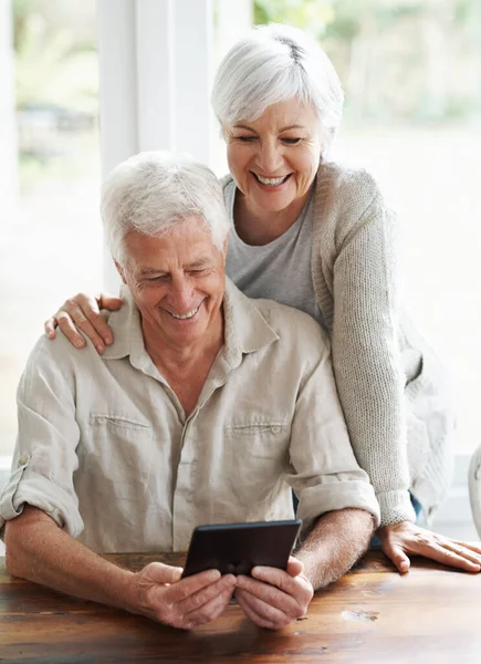 Olhe para essas fotos que os netos nos enviaram.... Um casal sênior examinando um tablet digital enquanto se sentam na mesa da sala de jantar. — Fotografia de Stock