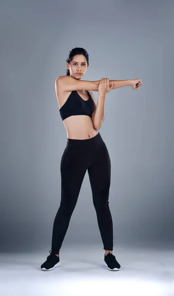Sträck ut innan den verkliga träningen börjar. Full längd skott av en sportig ung kvinna som står och sträcker armen mot en grå bakgrund. — Stockfoto