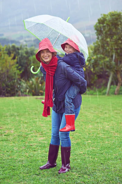 Brincalhão à chuva. Retrato recortado de uma mãe carregando seu filho de costas para fora na chuva. — Fotografia de Stock