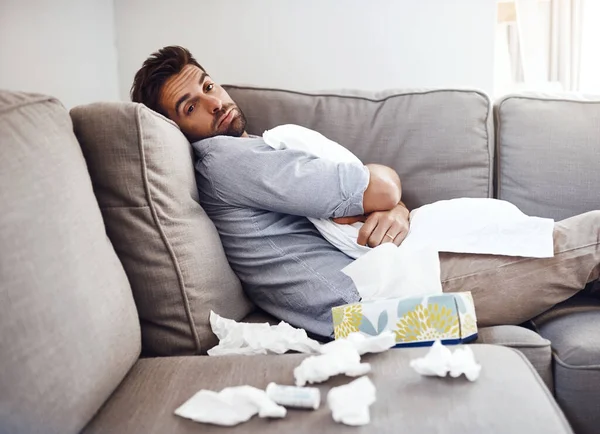 Anyone want to trade places. Portrait of a sickly young man holding a pillow while sitting on a couch with tissues laying all around at home. — Stock Photo, Image