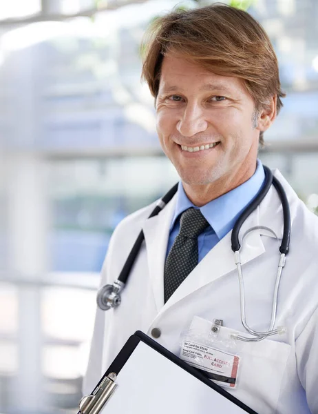 Meine Patienten stehen an erster Stelle. Ein hübscher Arzt lächelt in die Kamera, während er ein Klemmbrett hält. — Stockfoto