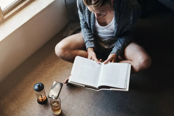 Passare un po 'di tempo da solo non e' sempre una cattiva idea. Colpo ad angolo alto di un giovane che legge un libro seduto sul pavimento di casa. — Foto Stock