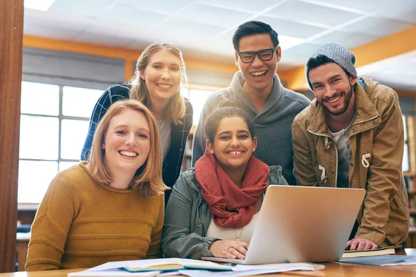 Het voelt een beetje beter als jullie samen studeren. Portret van een vrolijke jonge groep studenten die samenwerken met een laptop om te studeren voor examens in een bibliotheek. — Stockfoto