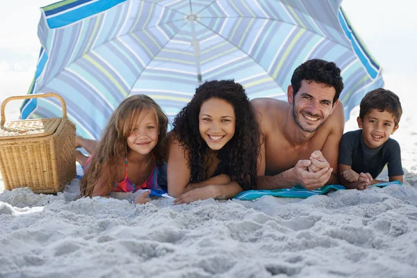 Enjoying a relaxing family vacation. A happy family smiling at the camera while lying under an umbrella at the beach. — Stock Photo, Image