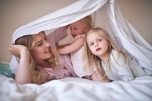 Las travesuras de tiempo de juego con las chicas. Tiro de una adorable familia de tres jugando bajo una sábana en la cama en casa. — Foto de Stock