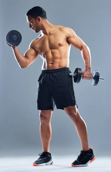 Det är mitt mål att fortsätta göra muskler. Studio skott av en muskulös ung man utövar med hantlar mot en grå bakgrund. — Stockfoto