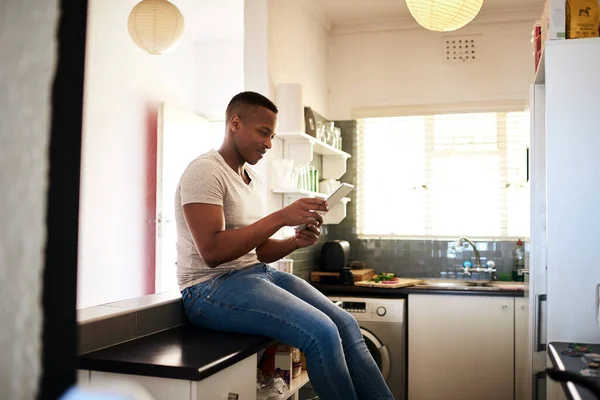 Internet parece interesante hoy en día. Recorte de un joven guapo usando una tableta digital mientras está sentado en el mostrador de la cocina en casa. —  Fotos de Stock