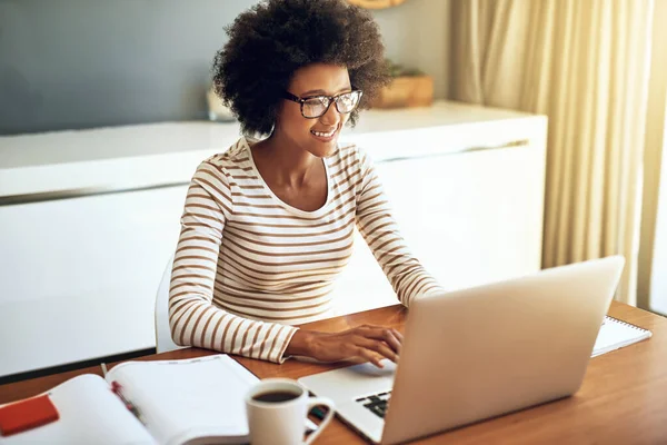 Ich bin am besten, wenn ich von zu Hause aus arbeite. Aufnahme einer selbstbewussten jungen Frau, die an ihrem Laptop arbeitet, während sie tagsüber zu Hause eine Tasse Kaffee trinkt. — Stockfoto