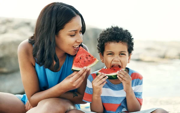 Watermelon is out favourite fruit. Shot of a mother and sitting down and enjoying some watermelon at the beach. — Stock Photo, Image