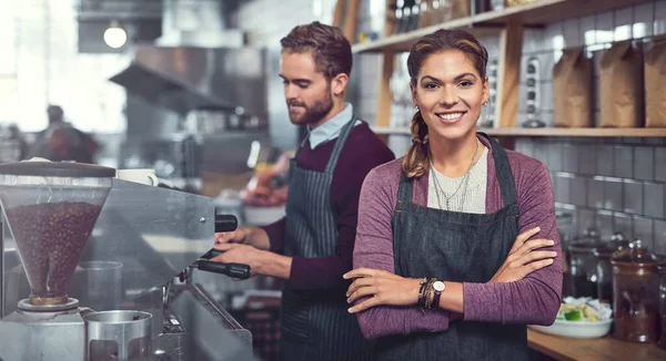 Starting a business - passion made visible. Portrait of a confident young woman working in a cafe while her coworker operates a coffee machine in the background.