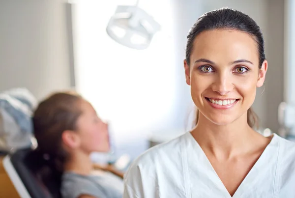 Sono felice di aver deciso di specializzarmi in odontoiatria. Una dentista donna in piedi nel suo studio con il suo paziente bambino sullo sfondo. — Foto Stock