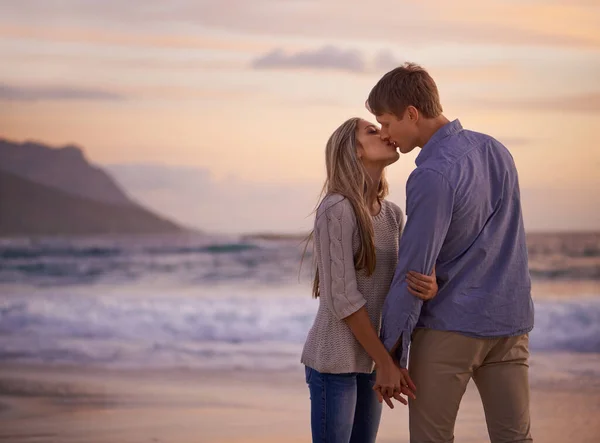 Varje kyss känns som vår första. Skjuten av ett ungt par njuter av en romantisk kyss på stranden vid solnedgången. — Stockfoto