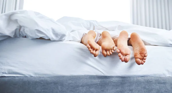 We all need a lazy day now and then. Rear view shot of a couples bare feet sticking out underneath the bedsheets at home. — Stock Photo, Image