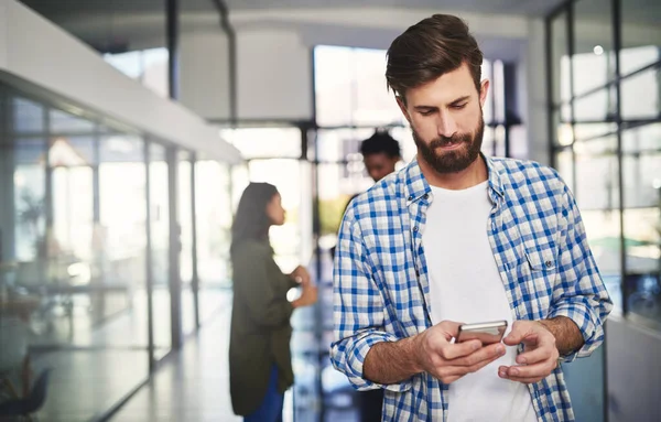 Posílání zpráv klientovi s nejnovějšími informacemi. Vystřižený snímek mladého návrháře pomocí mobilního telefonu v kanceláři s kolegy v pozadí. — Stock fotografie