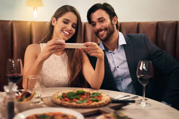 Quero lembrar-me desta pizza. Tiro de um jovem casal feliz tirando uma foto de sua refeição em um celular durante um jantar romântico em um restaurante. — Fotografia de Stock