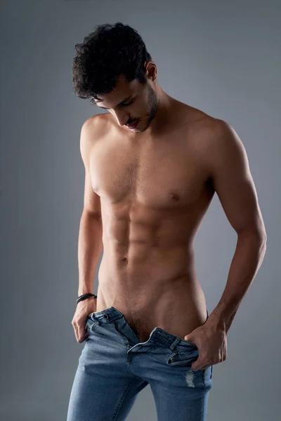 The perfect physique for a pair of jeans. Studio shot of a handsome young shirtless man undressing against a grey background. — Stock Photo, Image