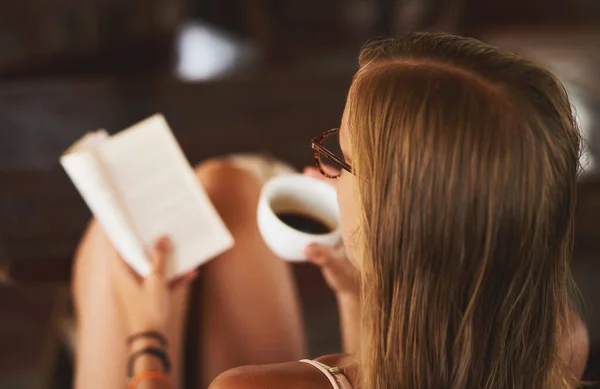 E 'il suo comfort preferito leggere. Foto posteriore di una giovane donna che legge un libro mentre beve una tazza di caffè. — Foto Stock