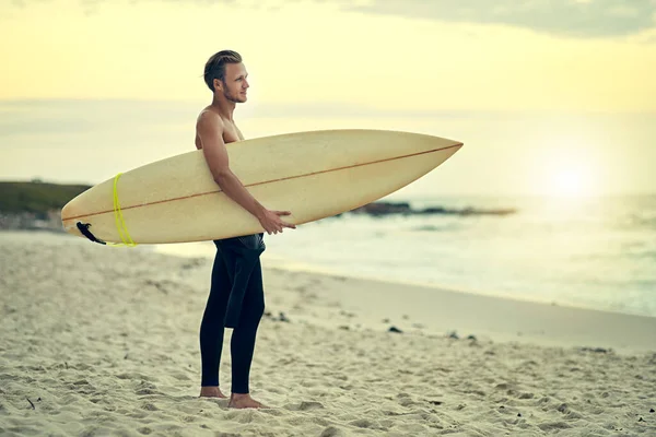Surfar não é um hobby, é um estilo de vida. Tiro de um jovem surfista sem camisa observando as ondas enquanto segurava sua prancha na praia. — Fotografia de Stock