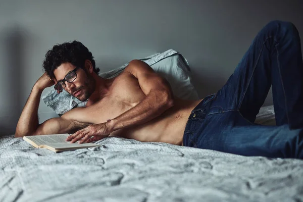 Ele gosta de ler romances no fim de semana. Tiro cortado de um belo jovem sem camisa lendo um livro em sua cama em casa. — Fotografia de Stock