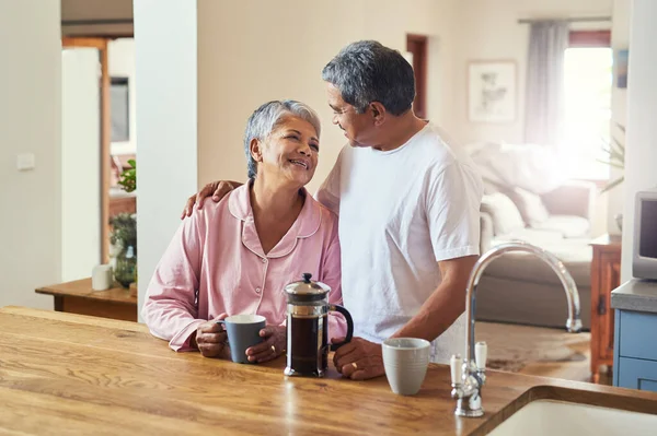 Empecemos bien por la mañana. Toma de una alegre pareja de ancianos abrazándose mientras se miran a los ojos y beben café en casa durante el día. — Foto de Stock