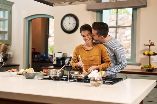 Te quiero, mi dulce tarta. Disparo de un joven cariñoso besando a su esposa en la mejilla mientras ella prepara una comida en la cocina en casa. — Foto de Stock