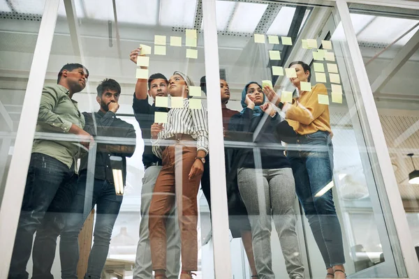 Sie arbeiten sich an ihren besten Konzepten ab. Aufnahme einer Gruppe von Geschäftsleuten beim Brainstorming mit Zetteln an einer Glaswand in einem Büro. — Stockfoto