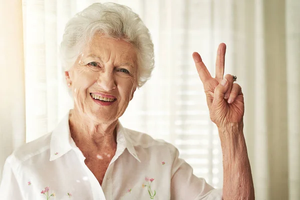 V jako vitalita. Portrét šťastné starší ženy ukazující mírové gesto doma. — Stock fotografie