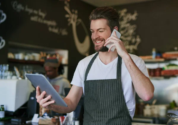 Just calling to let you know your order is ready. Shot of a young barista taking orders via cellphone and logging them on his tablet. — Stock Photo, Image