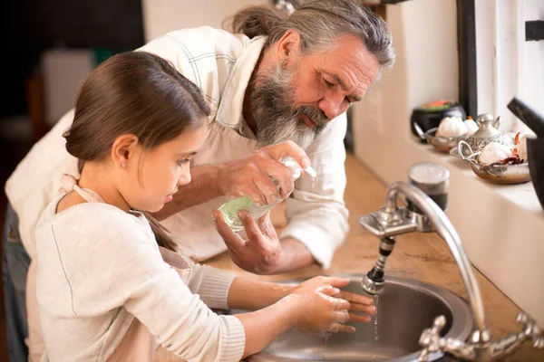 A lavar os germes. Tiro de uma menina lavando as mãos na pia da cozinha como seu avô está por perto. — Fotografia de Stock
