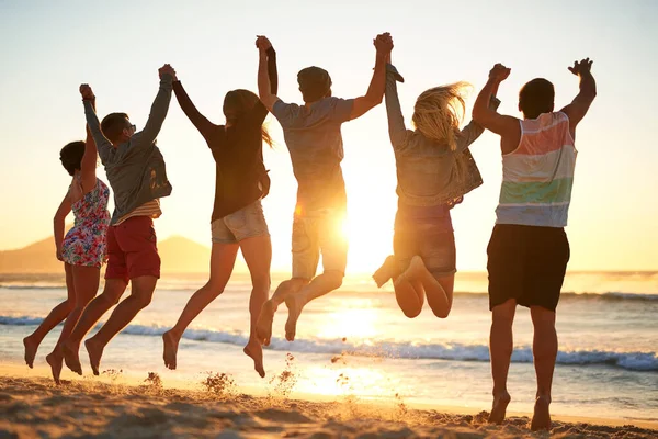 Yhdessä lennämme. Taustakuva joukosta nuoria ystäviä hyppäämässä ilmaan pitäen toisiaan kädestä rannalla.. — kuvapankkivalokuva