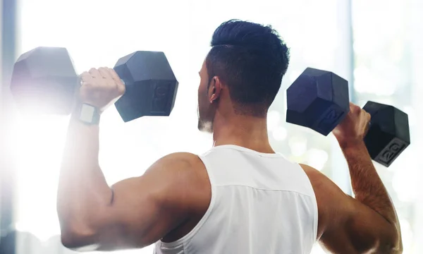 Det är ett åtagande han gör varje dag. Rearview bild av en stilig ung idrottsman som tränar med hantlar i ett gym. — Stockfoto