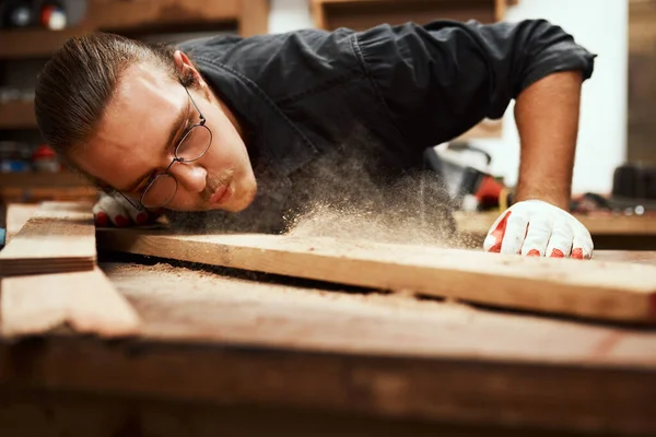 Det ser bra ut. Beskuren skott av en fokuserad ung man snickare blåser damm av en bit trä efter slipning den inne i sin verkstad under natten. — Stockfoto