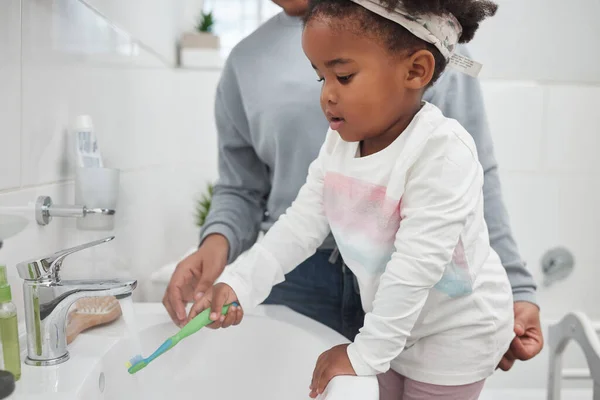 Lär dig allt om god hygien från ung ålder. Skjuten av en mor hjälpa sin lilla dotter skölja sin tandborste på en kran i badrummet hemma. — Stockfoto