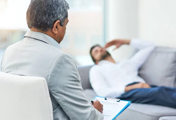 Relax and start from the beginning. Shot of an stressed out young man having a discussion with his doctor while lying on a sofa inside of a doctors office during the day. — ストック写真