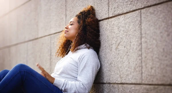 Elle a trouvé sa propre chanson. Tourné d'une jolie jeune femme assise et appuyée contre un bâtiment tout en écoutant de la musique dans la ville. — Photo
