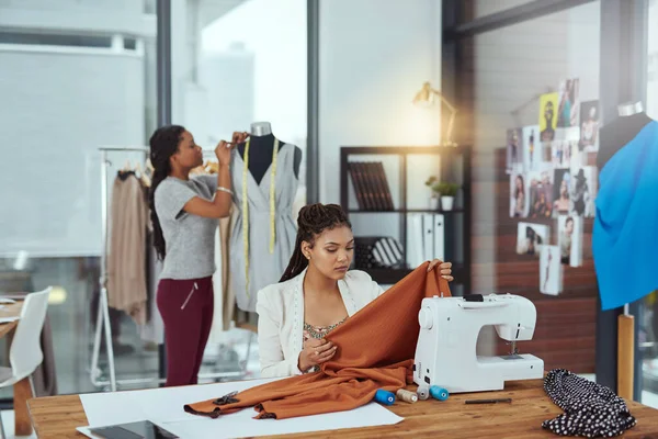 Sicherstellen, dass der Stoff von höchster Qualität ist. Aufnahme einer jungen Modedesignerin beim Nähen von Kleidungsstücken, während eine Kollegin im Hintergrund an einer Schaufensterpuppe arbeitet. — Stockfoto