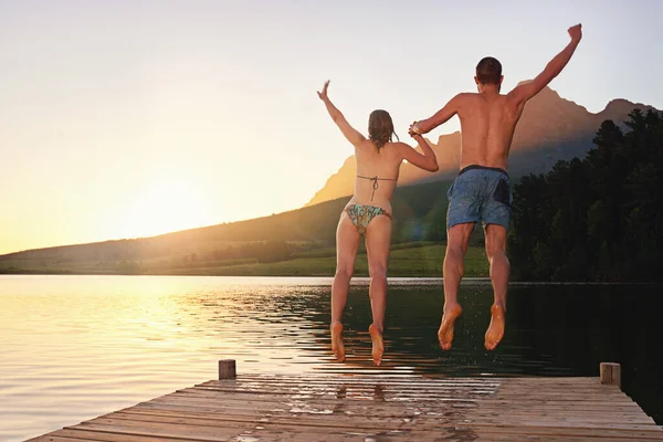 Taking the plunge. Rearview shot of a young couple in swimsuits leaping of a dock at sunset. — ストック写真
