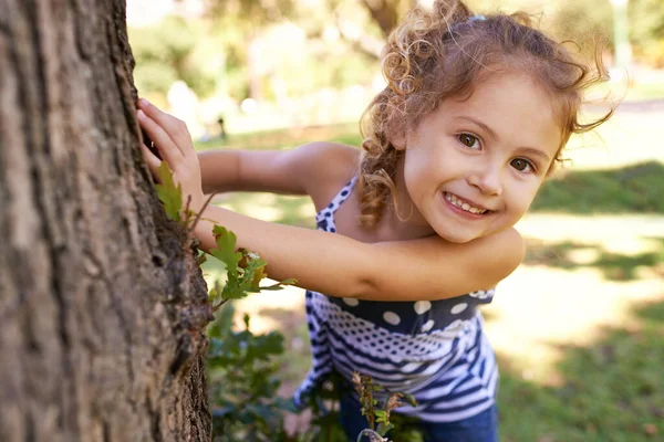 They wont find me here. Portrait of a little girl hiding behind a tree in a park. Stock Picture