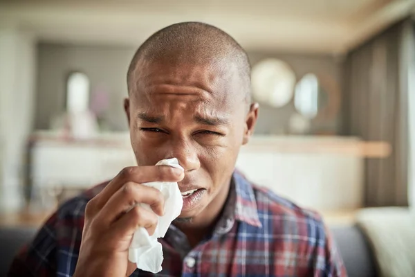 Im really not in the mood to go to work today. Portrait of an uncomfortable looking young man holding a tissue in front of his nose while being seated on a couch at home. — Stock Photo, Image