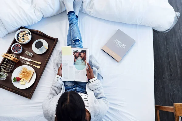 Frühstück im Bett und einige gute Bücher. Aufnahme einer unkenntlichen Frau, die eine Zeitschrift liest und zu Hause auf ihrem Bett frühstückt. — Stockfoto