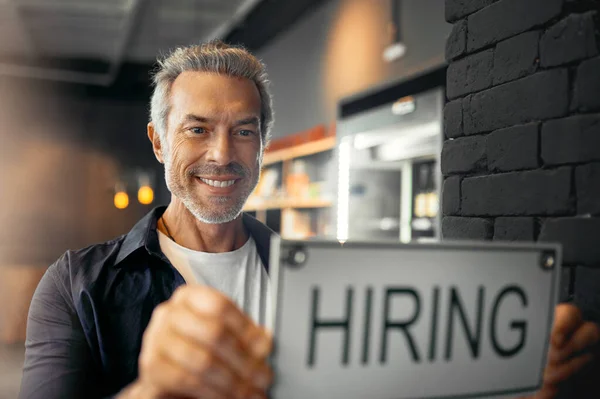 Estoy buscando nuevos miembros del personal. Retrato recortado de un apuesto hombre maduro sosteniendo un cartel de contratación mientras estaba de pie en su cafetería. — Foto de Stock