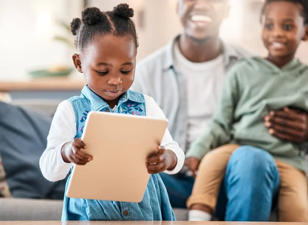 Het coolste vermaak om de kinderen tevreden te houden. Foto van een schattig klein meisje met behulp van een digitale tablet met haar familie op de achtergrond thuis. — Stockfoto