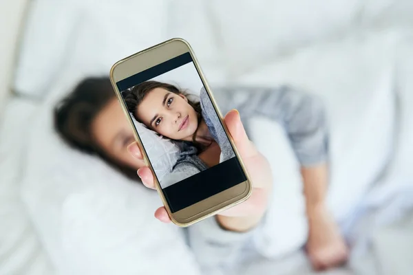 Selfies tôt le matin. Prise de vue d'une jolie jeune femme prenant un autoportrait avec son téléphone portable alors qu'elle était couchée dans son lit à la maison. — Photo